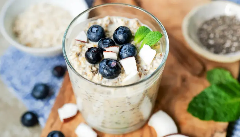 Ein Glas mit Overnight Oats Grundrezept, garniert mit Blaubeeren, Kokosraspeln und Minze. Daneben Chia-Samen und Haferflocken.