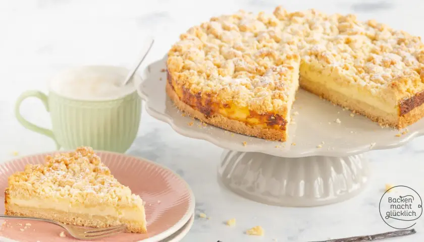 Ein saftiger Streuselkuchen mit Puddingfüllung, bestäubt mit Puderzucker, daneben eine Tasse Cappuccino.