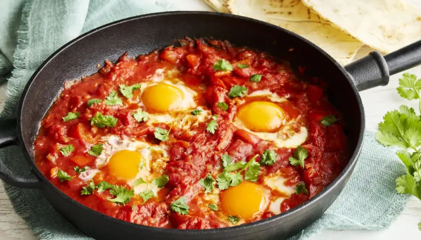 Herzhaftes Shakshuka Rezept in einer Gusseisenpfanne, mit vier pochierten Eiern in würziger Tomaten-Paprika-Sauce, garniert mit frischem Koriander.