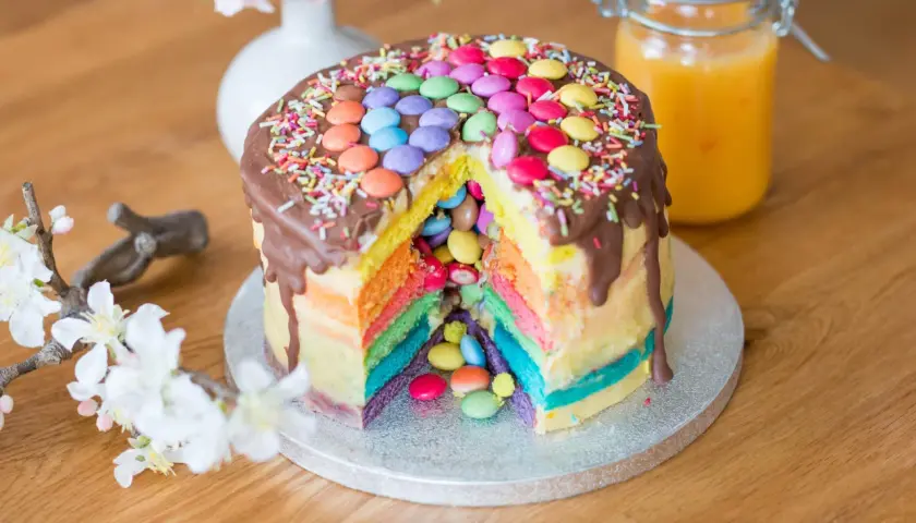 Bunter Smarties Kuchen mit Schokoladenglasur und Streuseln, ein Stück herausgeschnitten, das die Regenbogen-Schichten und Smarties im Inneren zeigt.