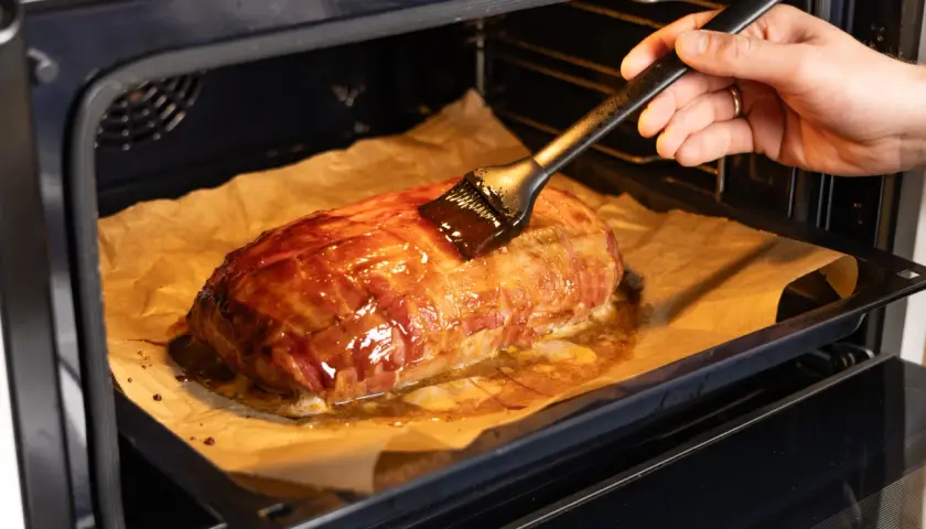 Ein Braten, umwickelt mit Bacon, wird im Backofen mit einer Glasur bestrichen. Die Bacon-Bomb-Zubereitung im Ofen ist fast fertig.
