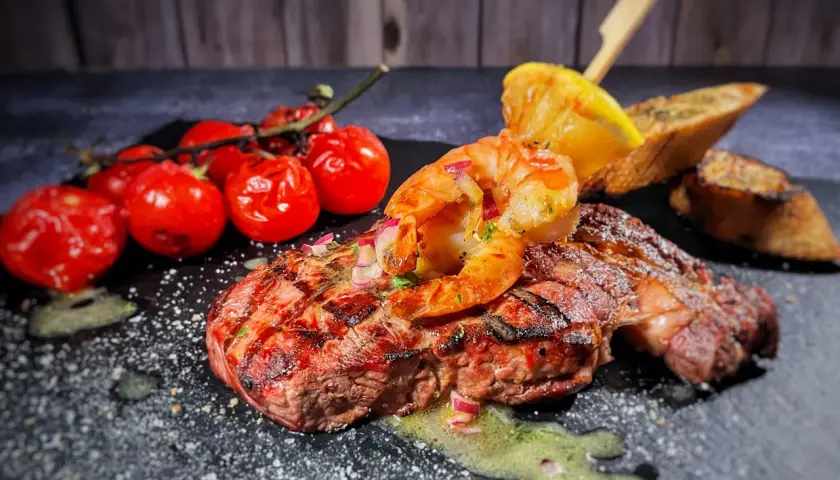 Perfektes Surf and Turf Rezept: saftiges Steak mit gegrillten Garnelen, Kirschtomaten, Zitrone und Brot auf dunklem Stein serviert.