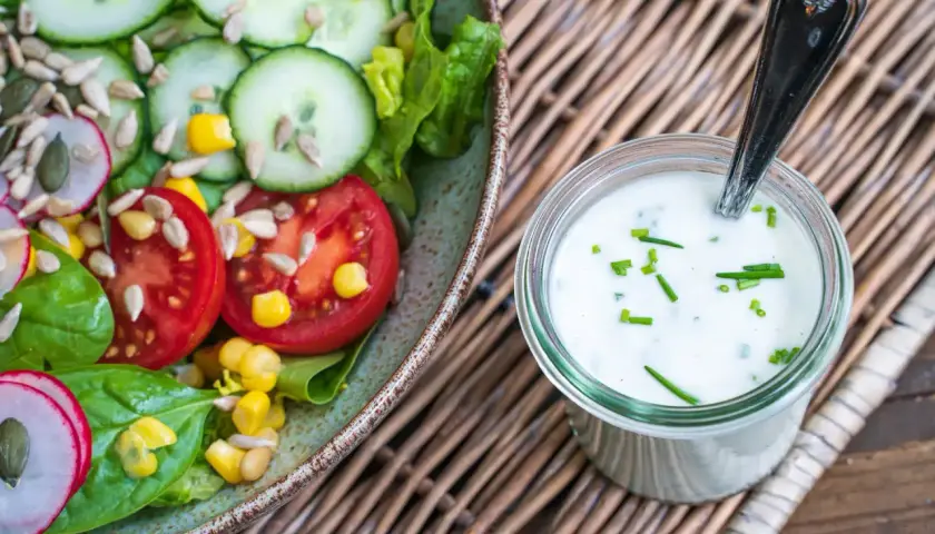 Frischer Salat mit Tomaten, Gurken und Radieschen neben einem Glas mit selbstgemachtem Joghurt-Dressing, garniert mit Schnittlauch.