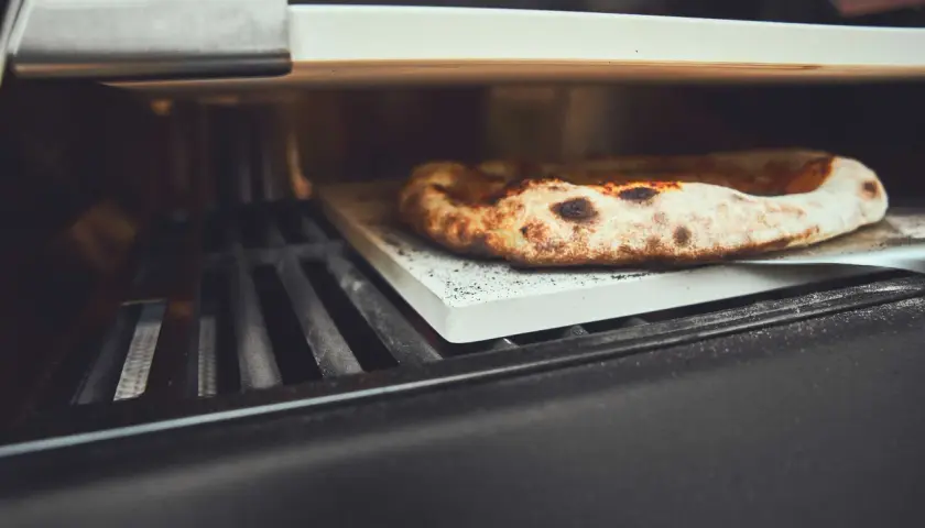 Eine köstliche Pizza wird auf einem Pizzastein im Gasgrill gebacken. Der Rand ist goldbraun und leicht angekohlt.