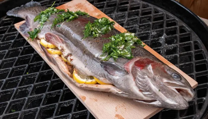 Eine ganze Forelle, gefüllt mit Zitronenscheiben und Kräutern, liegt auf einem Holzbrett auf dem Grillrost. Perfekt zum Forelle grillen!