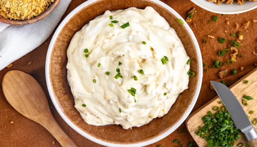 Cremiger Zwiebeldip, der an eine herzhafte Zwiebelsuppe erinnert, garniert mit frischen Schnittlauchstückchen. Daneben liegen knusprige Zwiebeln und ein Holzbrett mit gehacktem Schnittlauch.