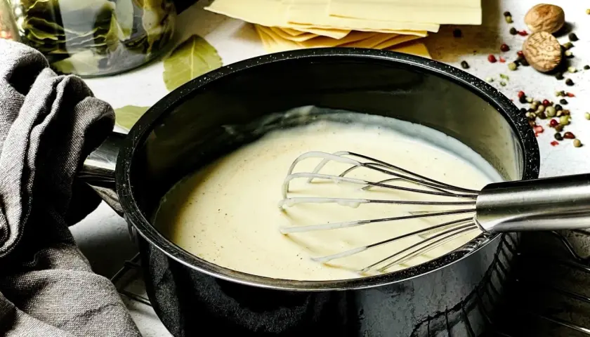 Cremige Béchamelsauce wird in einem Topf mit einem Schneebesen zubereitet. Daneben liegen Lasagneplatten und Gewürze.