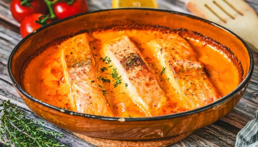 Saftige Lachsfilets in cremiger Lachs Sahne Sauce, garniert mit frischen Kräutern. Daneben Tomaten und eine Zitronenspalte.