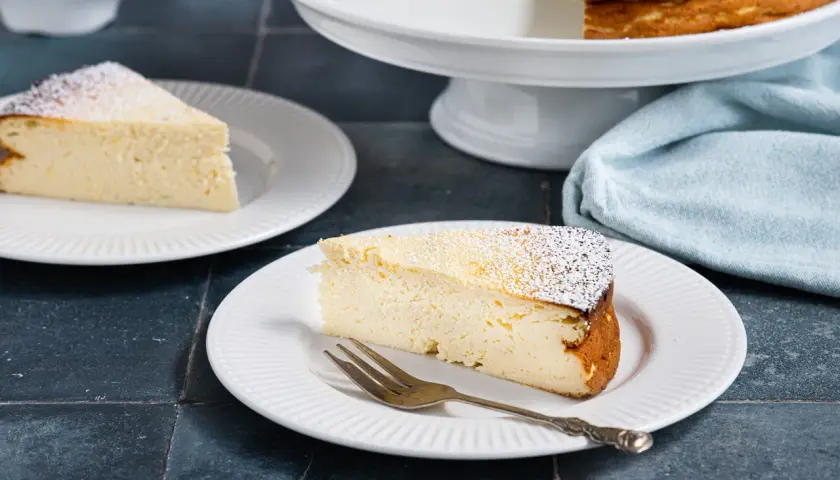 Ein saftiges Stück Quarkkuchen ohne Boden, bestäubt mit Puderzucker, auf einem Teller mit einer Gabel.