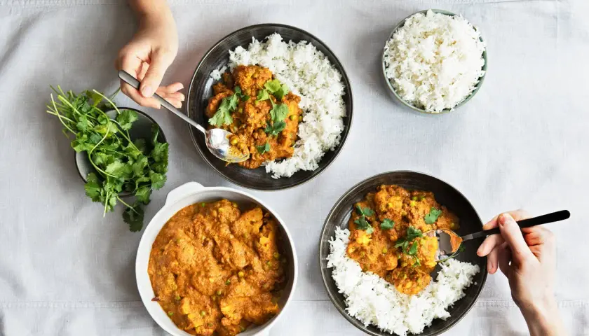 Drei Schalen mit Reis und leckeren indischen Gerichten, garniert mit Koriander. Eine Hand nimmt mit einem Löffel eine Portion Curry.