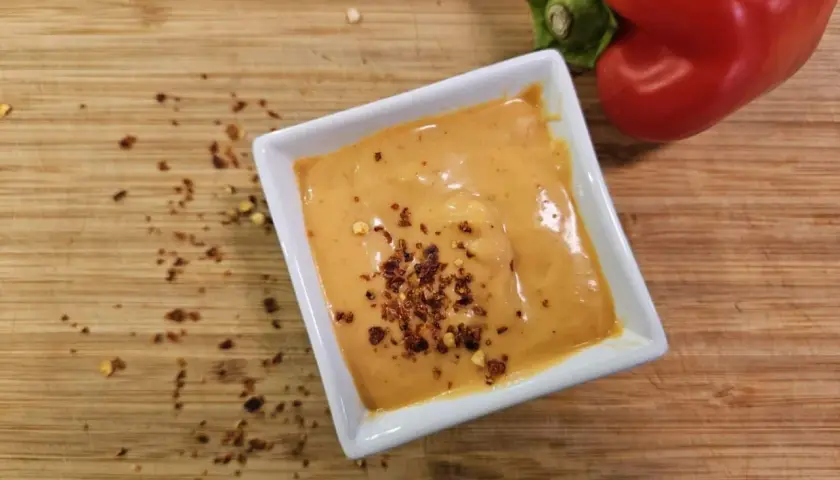 Cremige Chipotle-Sauce in einer weißen Schale, bestreut mit Chiliflocken. Daneben liegt eine rote Paprika auf einem Holzbrett.