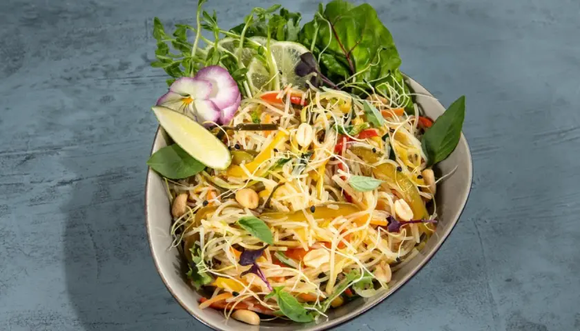 Bunter Asia Salat mit Reisnudeln, Erdnüssen, Gemüse und frischen Kräutern, garniert mit Limettenspalten und einer essbaren Blüte.