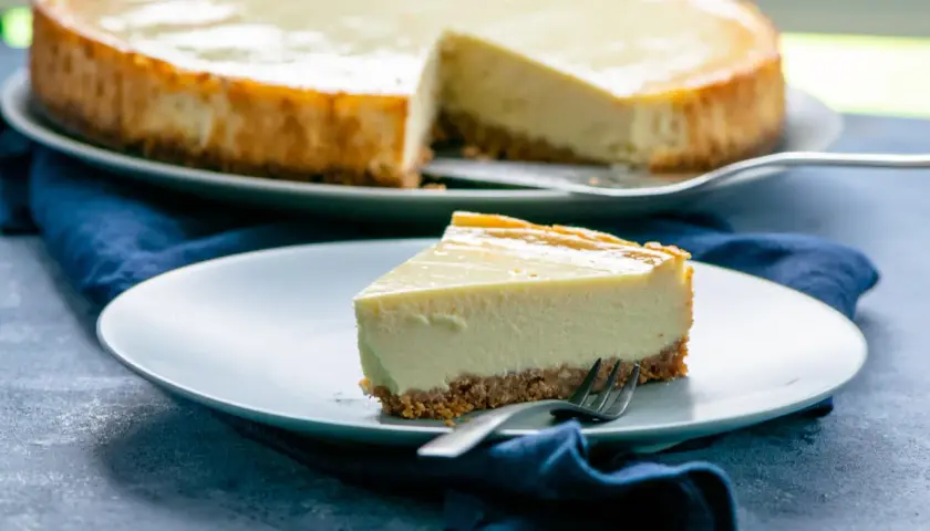 Ein Stück New York Cheesecake mit knusprigem Boden und cremiger Füllung auf einem Teller, daneben der ganze Kuchen.