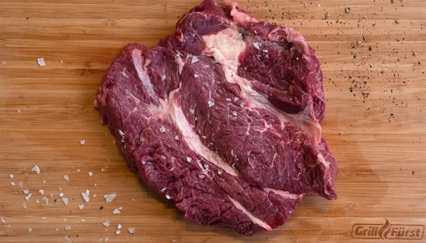 Ein roher Chuck Roast Braten, deutsch, liegt auf einem Holzbrett, bestreut mit grobem Salz und Pfeffer.