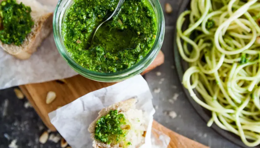 Frisches Basilikum-Pesto selber machen ist einfach! Hier ist ein Glas mit dem grünen Genuss, daneben Brot mit Pesto und eine Portion Spaghetti.