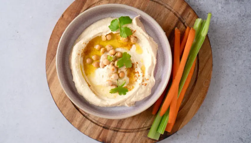 Cremiger Hummus mit Kichererbsen, Olivenöl und Petersilie, serviert mit Karotten- und Selleriesticks. Perfekt für ein schnelles Hummus Rezept.