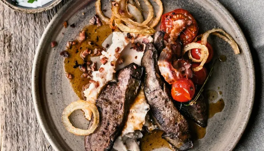 Herzhaftes leber braten mit Kartoffelpüree, Speck, Kirschtomaten und Röstzwiebeln auf einem rustikalen Teller.