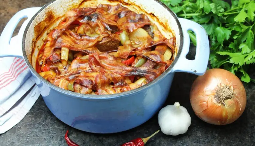 Herzhaftes Schichtfleisch im Backofen, bedeckt mit knusprigem Speck, daneben Zwiebel, Knoblauch und Chili.