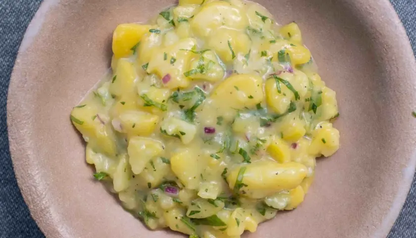 Ein cremiger Kartoffelsalat mit Speck, verfeinert mit frischer Petersilie und roten Zwiebeln, serviert in einer braunen Schale.