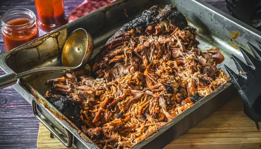 Saftiges pulled pork, frisch aus dem Gasgrill, in einer Metallschale mit Sauce und Fleischgabel.