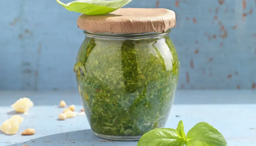 Frisches Pesto im Glas, garniert mit Basilikumblatt. Perfekt für dein nächstes Pesto Rezept. Pinienkerne und Parmesan liegen daneben.