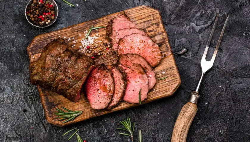 Saftiges Roastbeef, perfekt gegrillt und in Scheiben geschnitten, liegt auf einem Holzbrett. Gewürze und Rosmarin garnieren das Gericht.