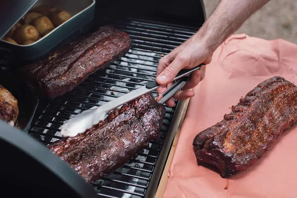 Spareribs werden mit Zangen auf dem Grill gewendet. Daneben liegen weitere Rippchen auf Butcher Paper.