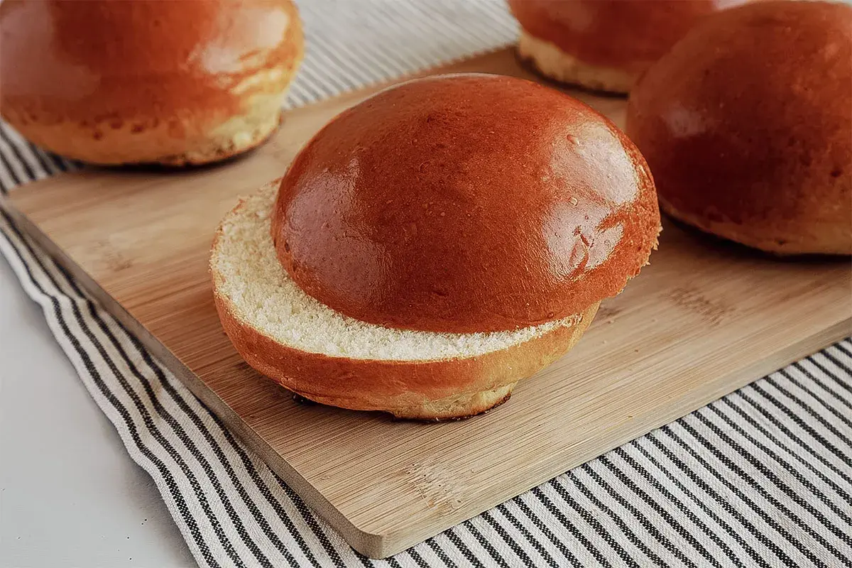 Ein aufgeschnittener, goldbrauner Brioche-Bun liegt auf einem Holzbrett. Weitere Buns sind im Hintergrund zu sehen.