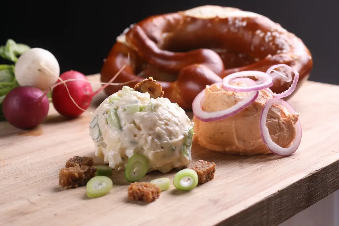 Zwei Portionen Obazda, garniert mit Zwiebelringen und Kräutern, daneben Radieschen und ein Brezel. Perfekt für ein Obazda Rezept!