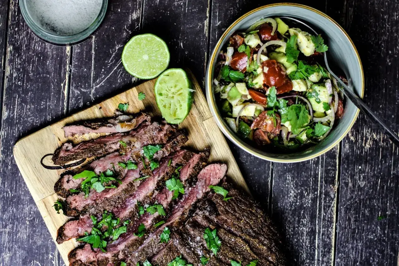 Perfekt gegrilltes Flanksteak, in Scheiben geschnitten, mit frischem Koriander und Limettenspalten, daneben ein bunter Salat.