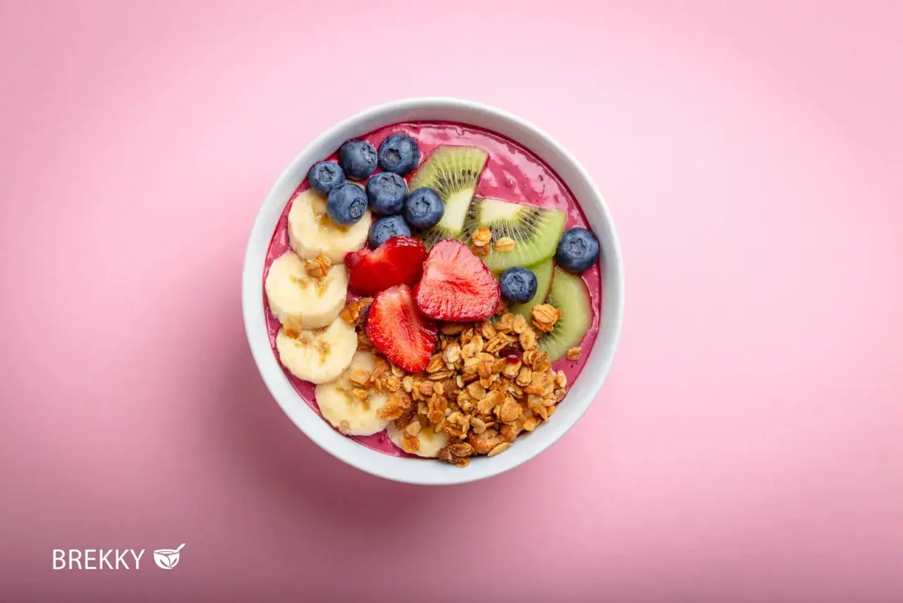 Ein farbenfrohes Acai Bowl Rezept mit Bananen, Erdbeeren, Blaubeeren, Kiwi und Granola auf rosa Hintergrund.