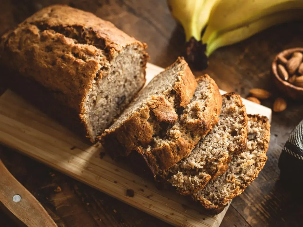 Ein saftiges Bananenbrot ohne Zucker, frisch aufgeschnitten auf einem Holzbrett. Daneben liegen reife Bananen und Mandeln.
