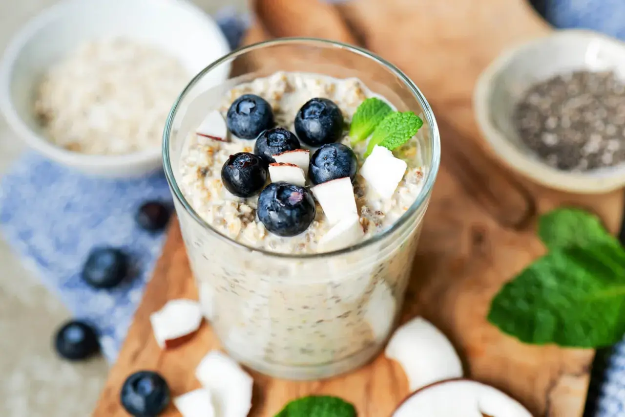 Ein Glas mit Overnight Oats Grundrezept, garniert mit Blaubeeren, Kokosraspeln und Minze. Daneben Chia-Samen und Haferflocken.