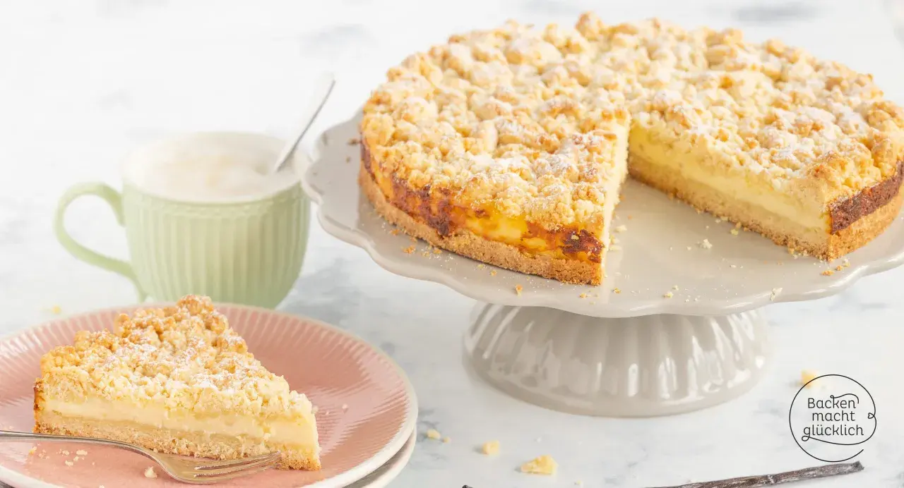 Ein saftiger Streuselkuchen mit Puddingfüllung, bestäubt mit Puderzucker, daneben eine Tasse Cappuccino.