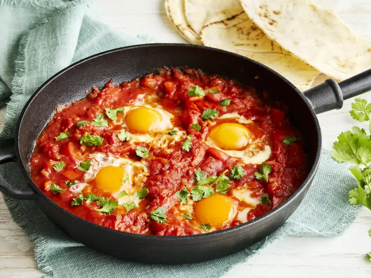 Herzhaftes Shakshuka Rezept in einer Gusseisenpfanne, mit vier pochierten Eiern in würziger Tomaten-Paprika-Sauce, garniert mit frischem Koriander.