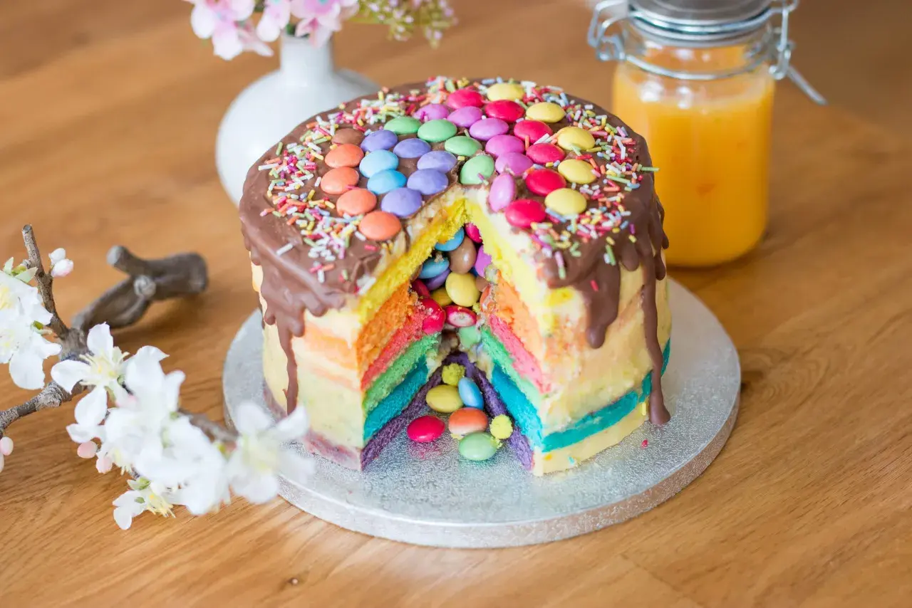 Bunter Smarties Kuchen mit Schokoladenglasur und Streuseln, ein Stück herausgeschnitten, das die Regenbogen-Schichten und Smarties im Inneren zeigt.