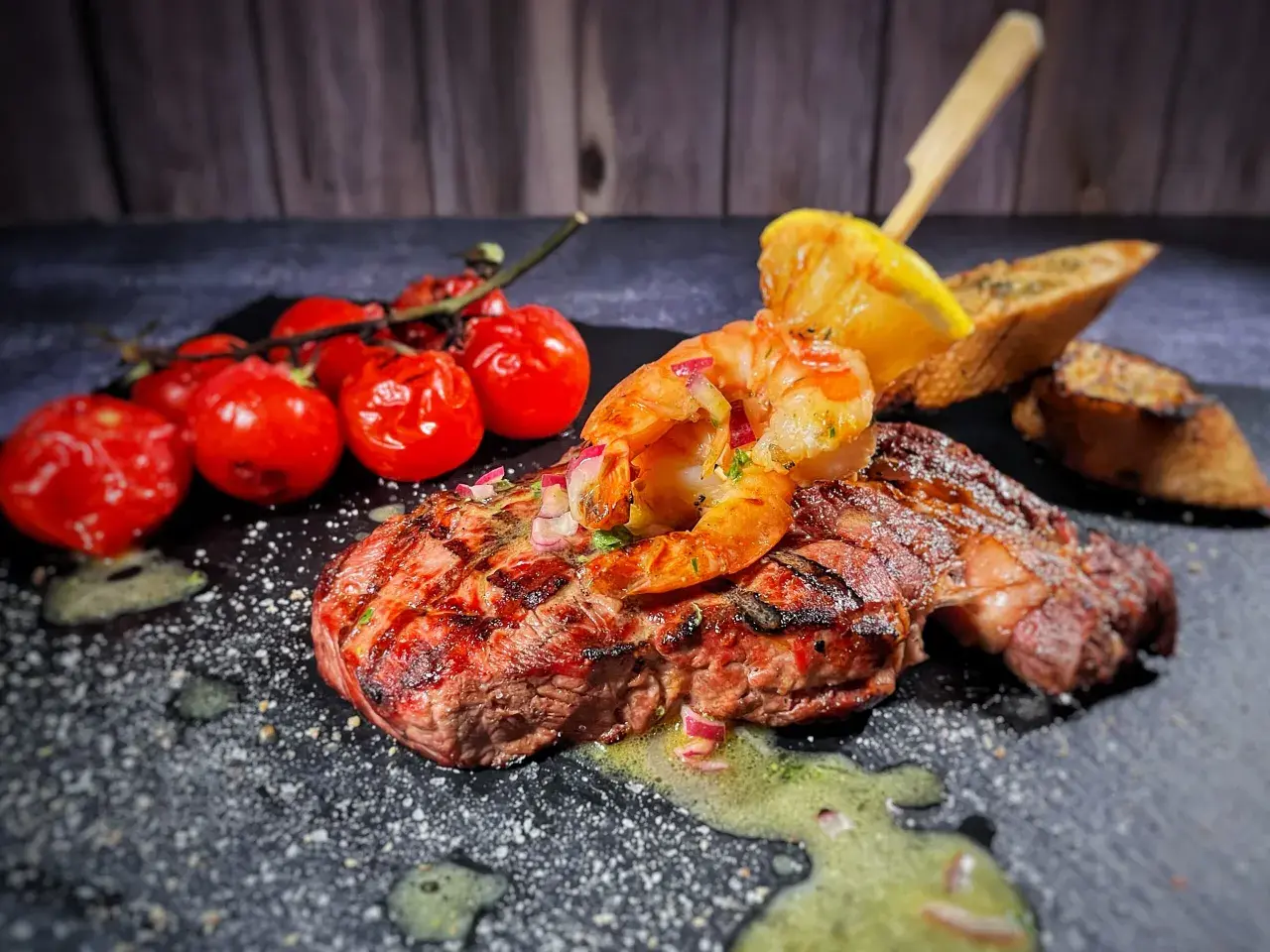 Perfektes Surf and Turf Rezept: saftiges Steak mit gegrillten Garnelen, Kirschtomaten, Zitrone und Brot auf dunklem Stein serviert.
