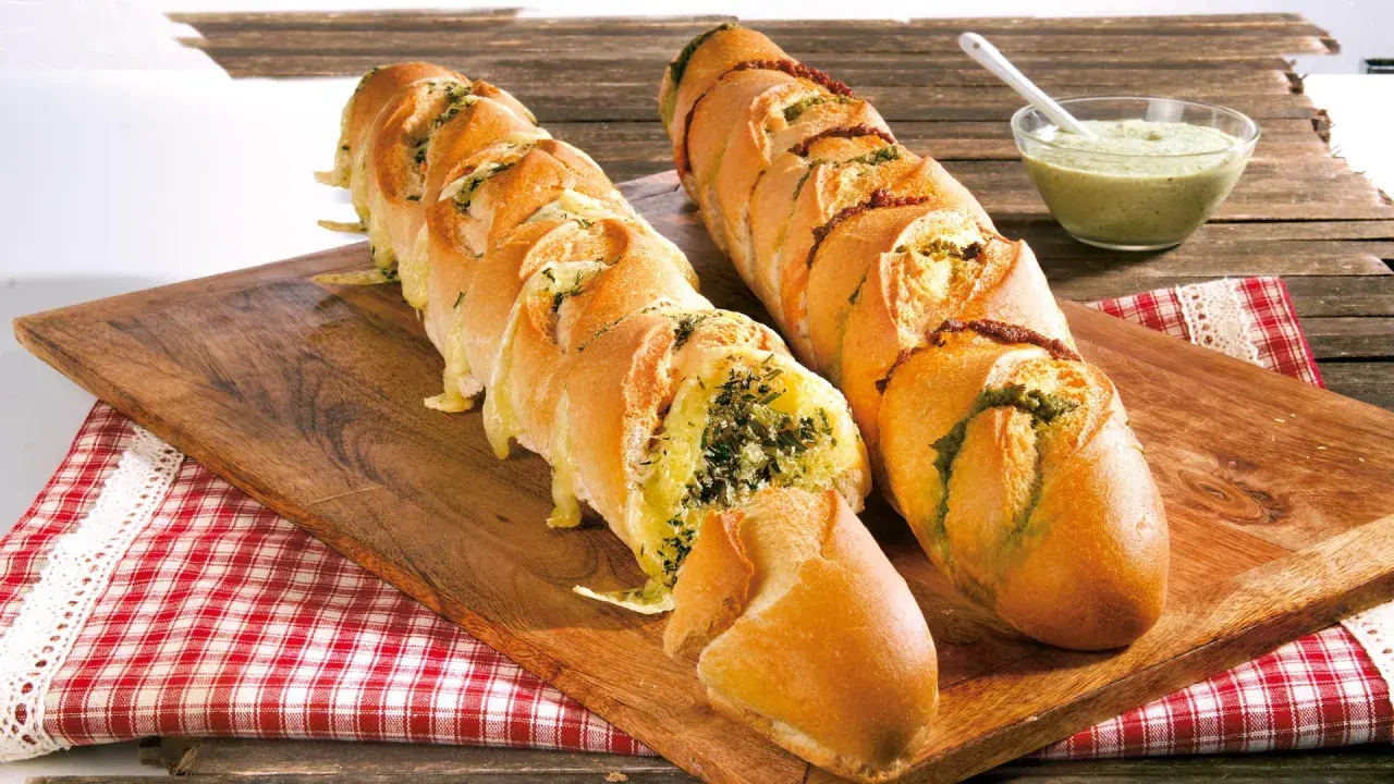Zwei knusprige Baguettes zum Grillen, gefüllt mit Kräutern und Käse, daneben eine Schale Dip.