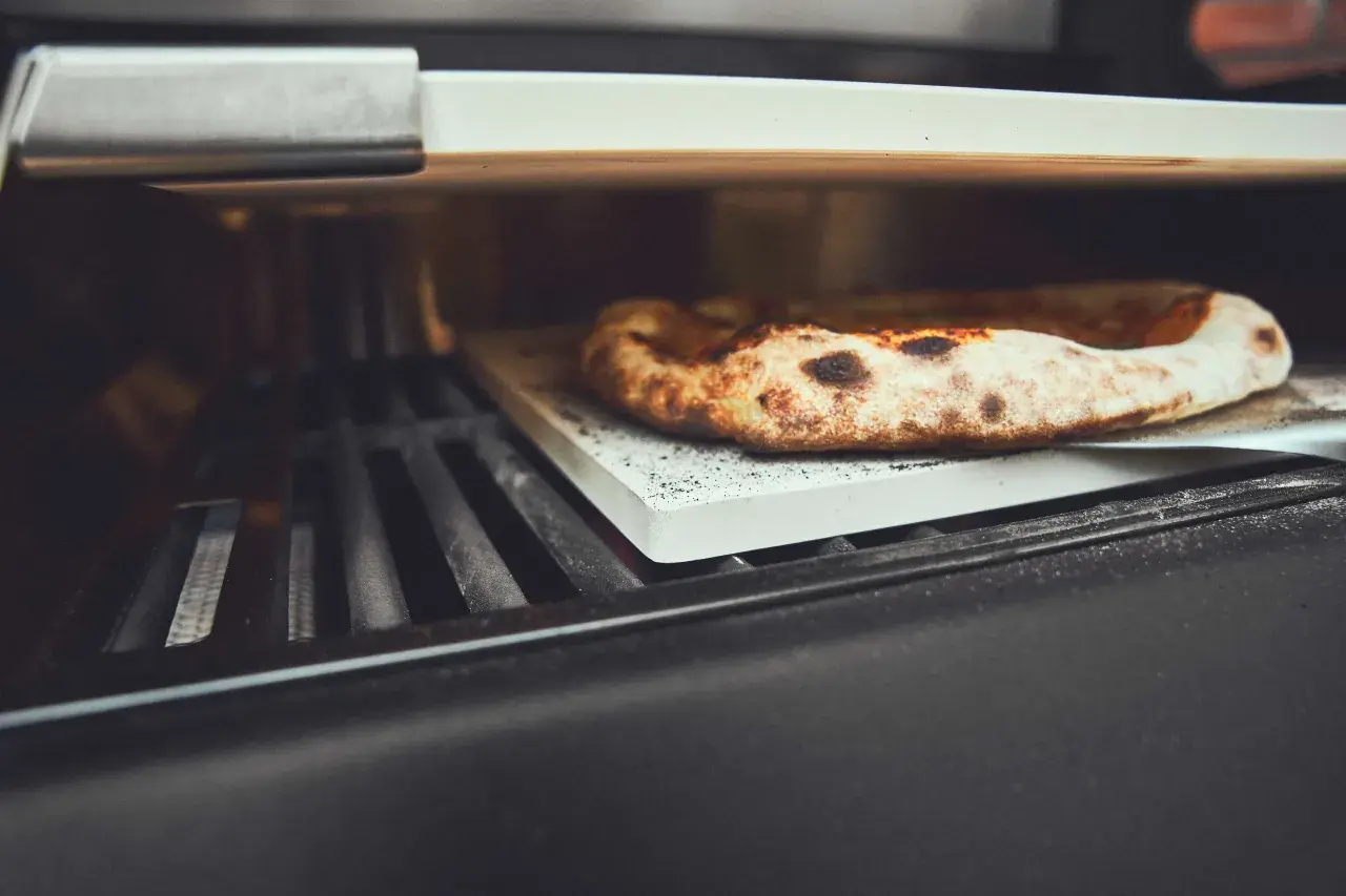 Eine köstliche Pizza wird auf einem Pizzastein im Gasgrill gebacken. Der Rand ist goldbraun und leicht angekohlt.