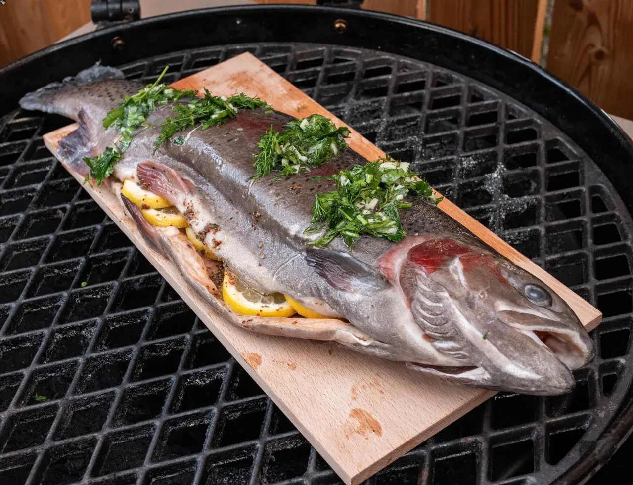 Eine ganze Forelle, gefüllt mit Zitronenscheiben und Kräutern, liegt auf einem Holzbrett auf dem Grillrost. Perfekt zum Forelle grillen!