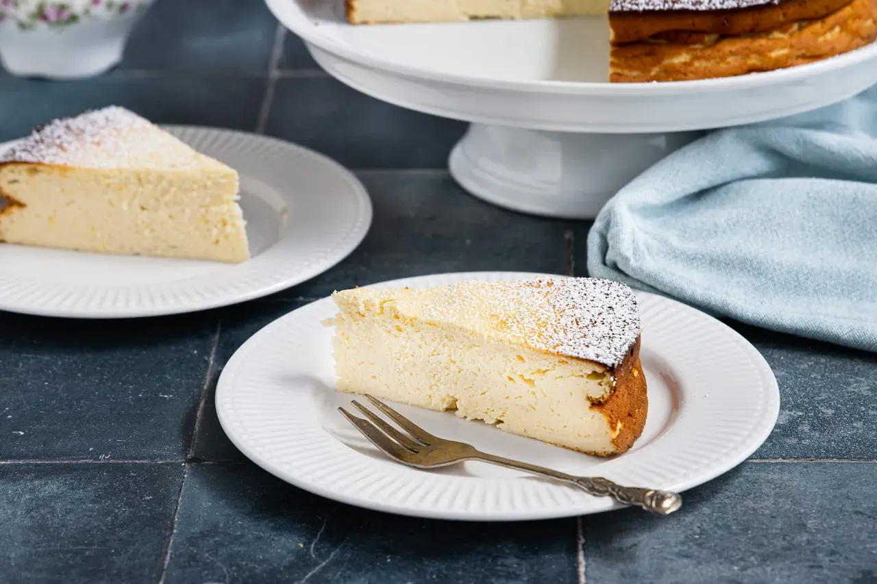 Ein saftiges Stück Quarkkuchen ohne Boden, bestäubt mit Puderzucker, auf einem Teller mit einer Gabel.