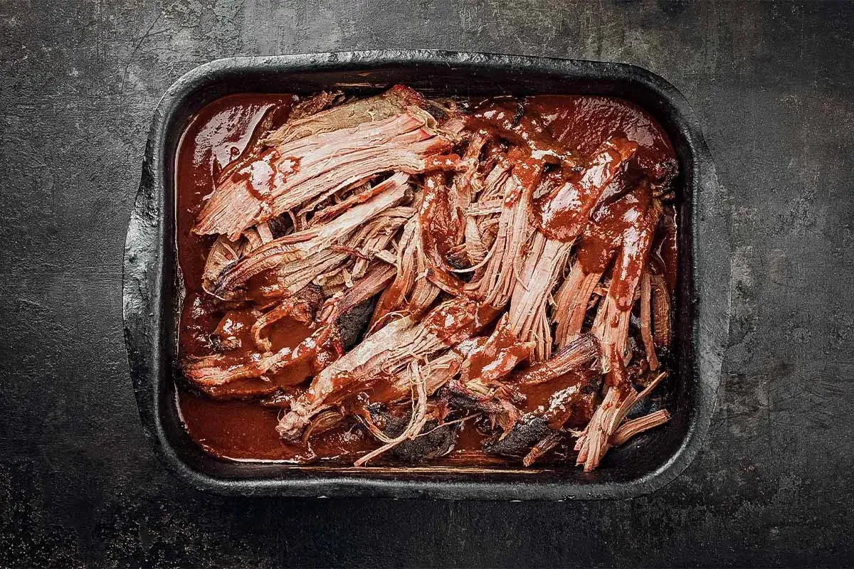 Saftiges pulled pork auf dem grill, in einer dunklen Schale mit BBQ-Sauce übergossen.
