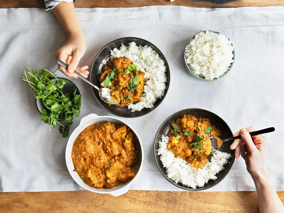Drei Schalen mit Reis und leckeren indischen Gerichten, garniert mit Koriander. Eine Hand nimmt mit einem Löffel eine Portion Curry.
