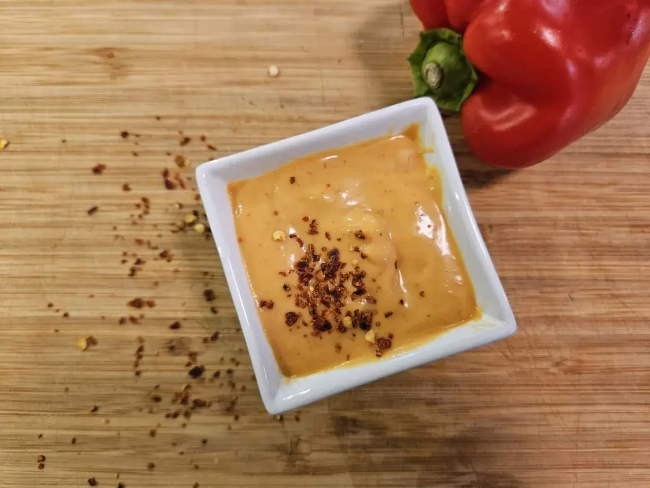 Cremige Chipotle-Sauce in einer weißen Schale, bestreut mit Chiliflocken. Daneben liegt eine rote Paprika auf einem Holzbrett.