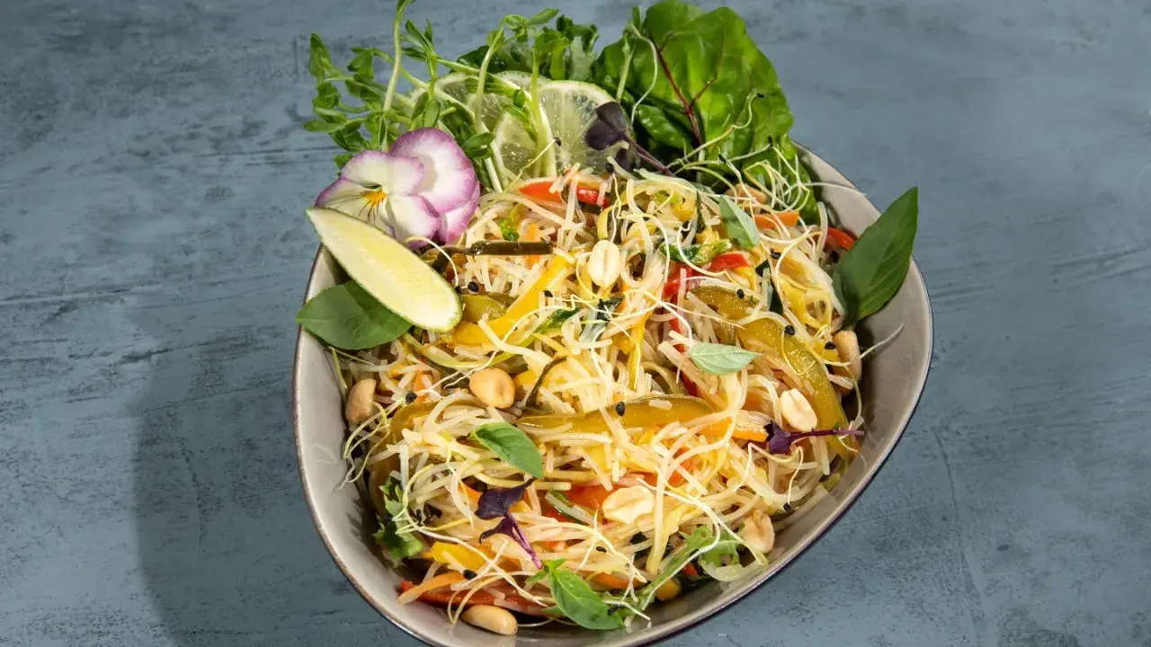 Bunter Asia Salat mit Reisnudeln, Erdnüssen, Gemüse und frischen Kräutern, garniert mit Limettenspalten und einer essbaren Blüte.