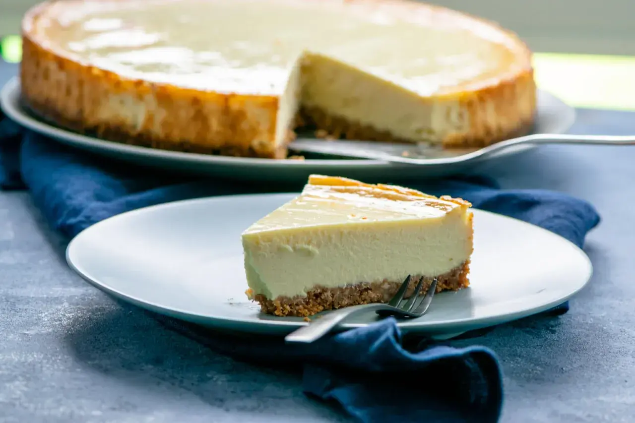 Ein Stück New York Cheesecake mit knusprigem Boden und cremiger Füllung auf einem Teller, daneben der ganze Kuchen.