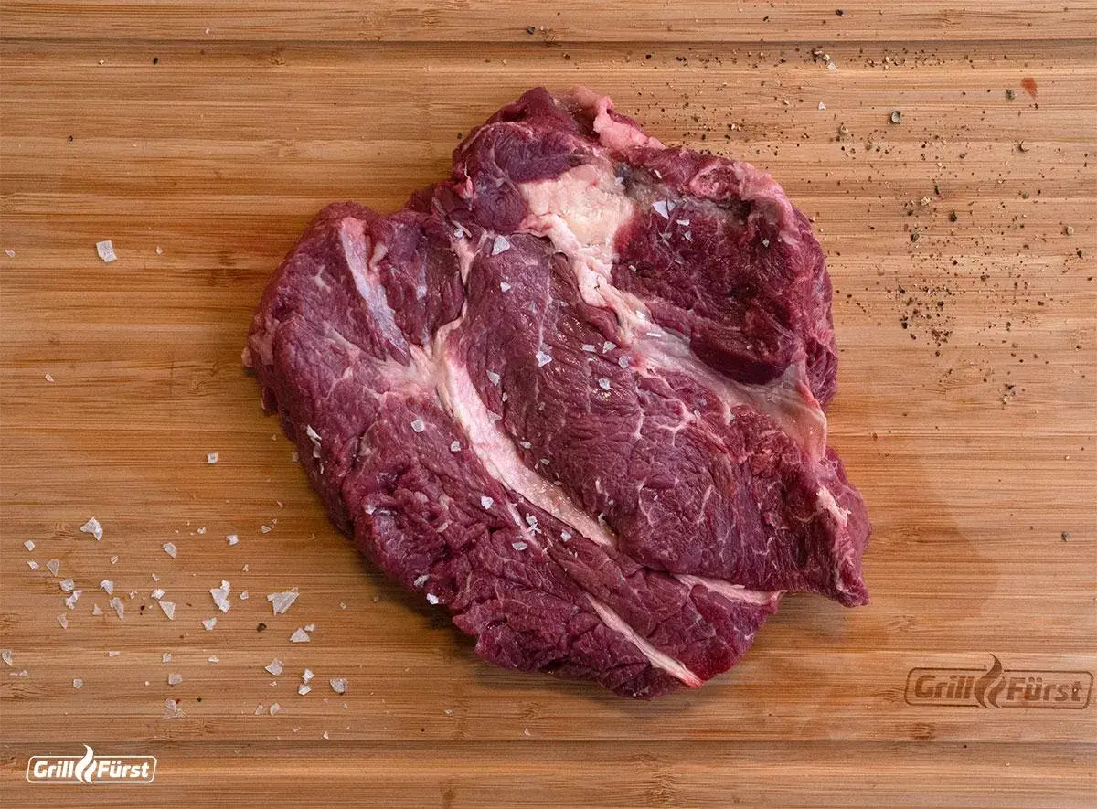 Ein roher Chuck Roast Braten, deutsch, liegt auf einem Holzbrett, bestreut mit grobem Salz und Pfeffer.