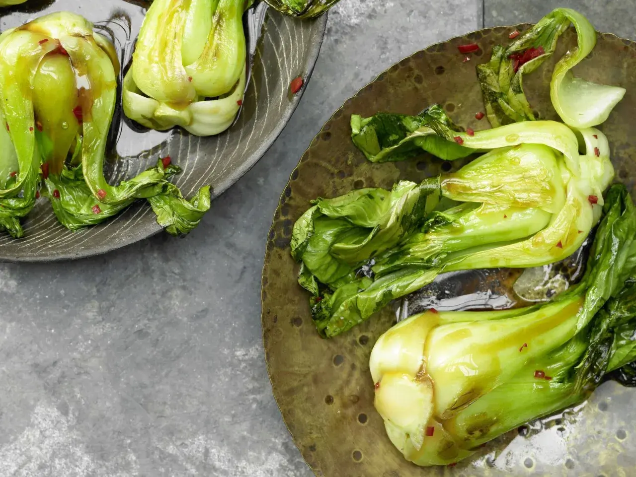 Zwei Teller mit gedünstetem Pak Choi, glänzend von einer Sauce und bestreut mit roten Chiliflocken.