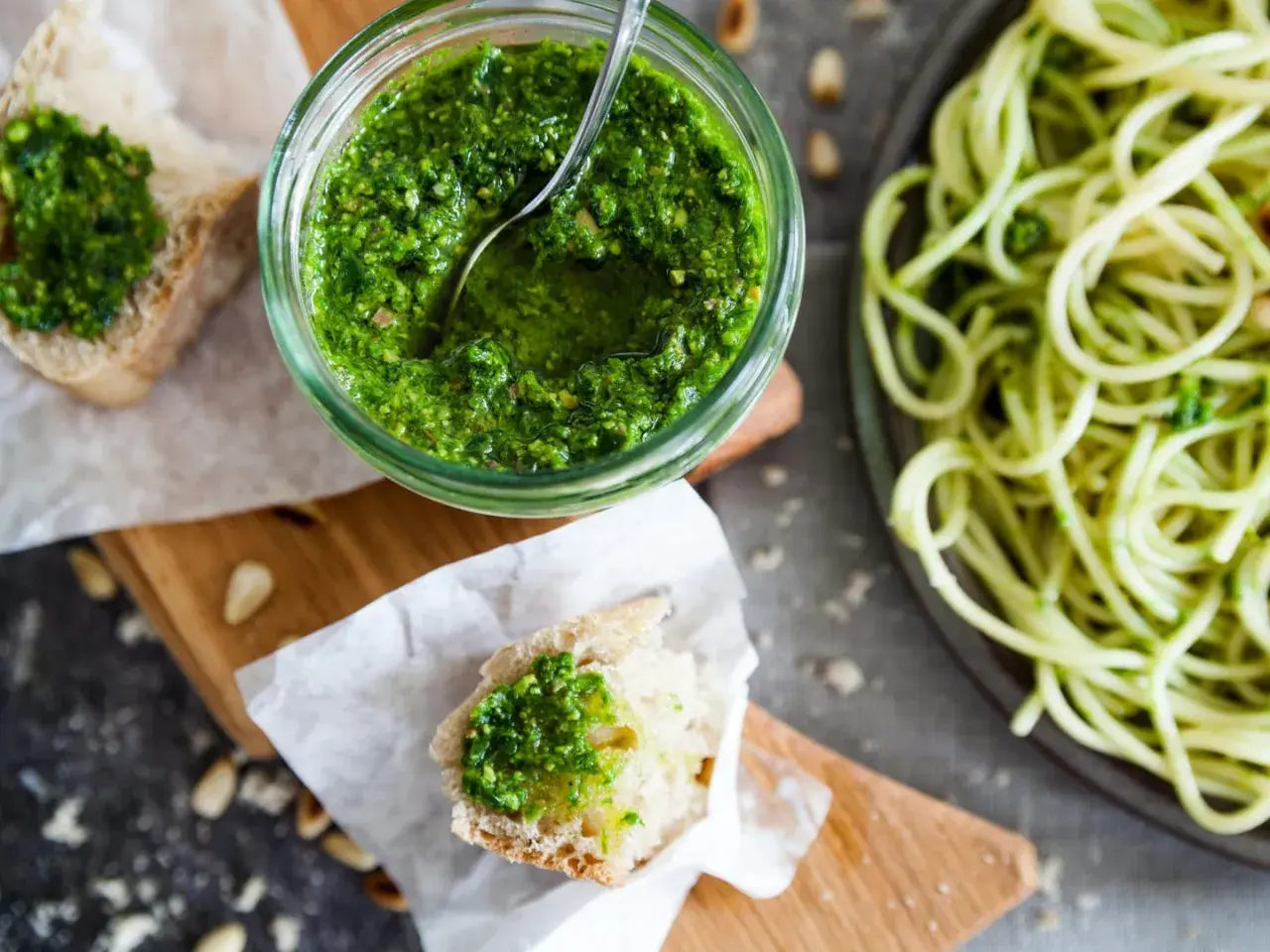 Frisches Basilikum-Pesto selber machen ist einfach! Hier ist ein Glas mit dem grünen Genuss, daneben Brot mit Pesto und eine Portion Spaghetti.