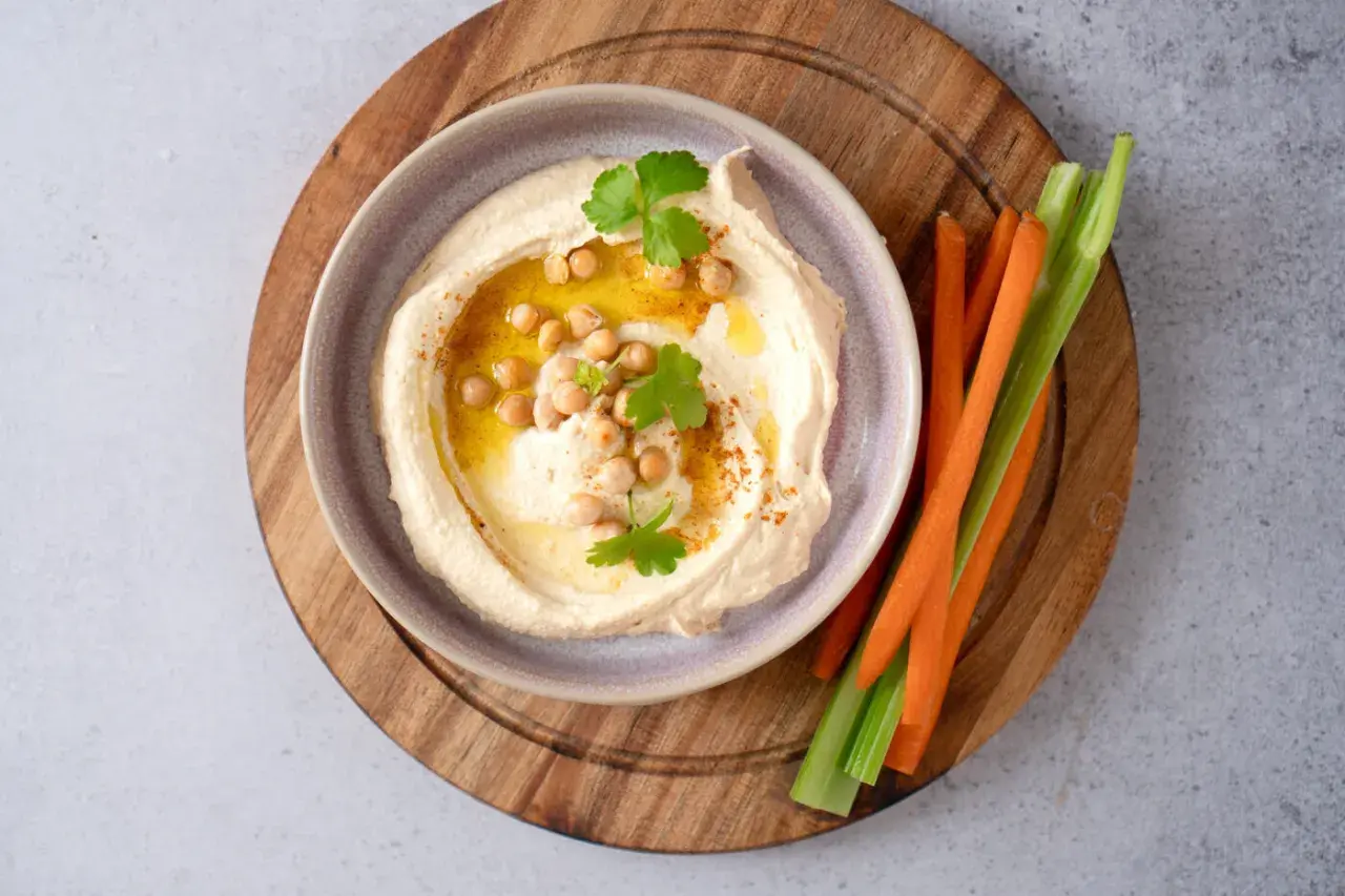 Cremiger Hummus mit Kichererbsen, Olivenöl und Petersilie, serviert mit Karotten- und Selleriesticks. Perfekt für ein schnelles Hummus Rezept.